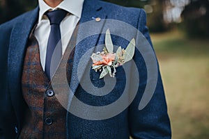 The groom in a suit and a vest stands in the woods, a buttonhole of flowers and eucalyptus on the groom`s jacket