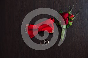 Groom wedding accessories. Red Boutonniere, gold rings and bow-tie on brown background