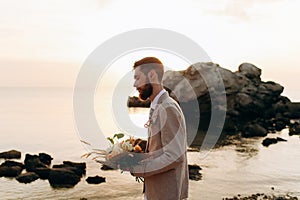 Groom with a wedding bouquet on the beach. Groom in boho style.