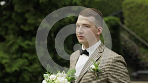 Groom, young man in the green summer park with a wedding bouquet. He waiting for his beloved bride