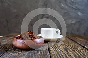 Ground coffee poured into a wooden bowl stands on a wooden table. White cup. Gray concrete wall