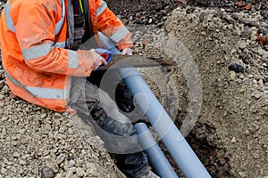 Groundworker in hi-viz coat cutting grey plastic telecom duct with hand saw while sitting