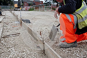 Groundworker in orange safety hi vis trousers fixing a timber along string line with steel pin to form a kerb riser