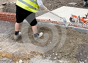 Groundworker placing semi-dry concrete around concrete edging kerb during footpath construction