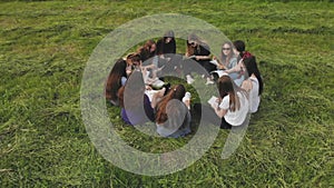 A group of 11 girls are sitting with notebooks on the grass in the park. Student work.