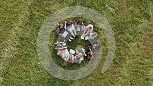 A group of 11 girls are sitting with notebooks on the grass in the park. Student work.