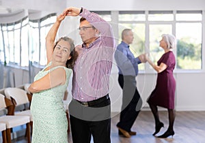 Group of active aged people rehearsing social dance in pair in dance hall