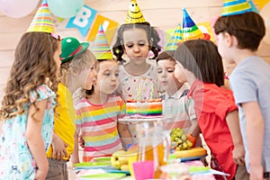 Group of adorable kids gathered around festival table. Children blow out candles on cake. Birthday party for