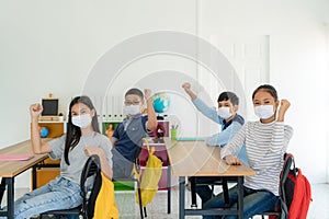 Group of Asian elementary school students wearing hygienic mask in classroom arm raised to be happy while back to school reopen