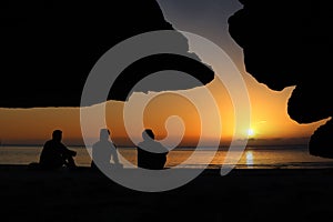 A group of Asian men sitting and looking at the sunset, beautiful red sky. In front of a cave on the beach