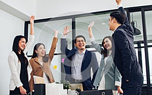 Group of Asian people wearing formal business suits standing together, raising hands with happiness, celebrating on success in