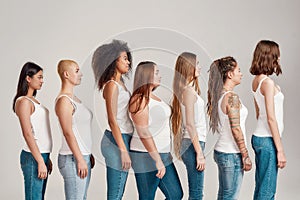 Group of beautiful diverse young women wearing white shirt and denim jeans looking aside while posing, standing isolated
