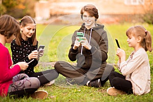 Group of busy kids looking at their phones texting sms and play games sitting with cross legs on grass outside in park