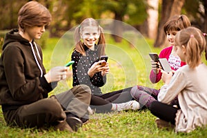 Group of busy kids looking at their phones texting sms and play games sitting with cross legs on grass outside in park