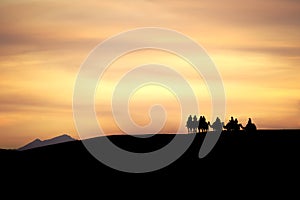 A group of camels and their riders arriving at the top of a sand dune are silhouetted against a dramatic cloudy desert sunset.