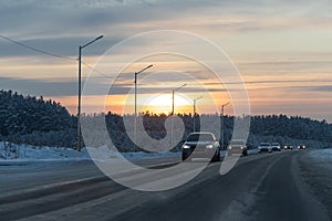 Group of cars with white lights is on the snowy road in winter on the orange sunset background