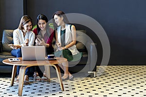 Group of charming beautiful Asian women using smartphone and laptop, chatting on sofa at cafe, modern lifestyle