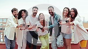 Group of cheerful best friends on a rooftop wine party. Crowd of young people drinking, toasting glasses, laughing and