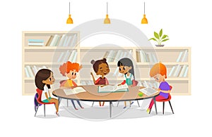 Group of children sitting around table at school library and listening to girl reading book out loud against bookcase or