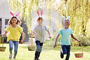 Group Of Children Wearing Bunny Ears Running On Easter Egg Hunt In Garden
