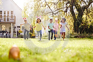 Group Of Children Wearing Bunny Ears Running To Pick Up Chocolate Egg On Easter Egg Hunt In Garden