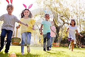 Group Of Children Wearing Bunny Ears Running To Pick Up Chocolate Egg On Easter Egg Hunt In Garden photo