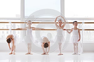 Group of children in white tutu training poses near barre in light room. Classical ballet school.