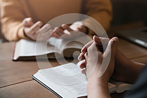 A group of Christians sit together and pray around a wooden table with blurred open Bible pages in their homeroom. Prayer for