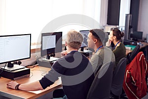 Group Of College Students Studying Computer Design Sitting At Line Of Monitors In Classroom