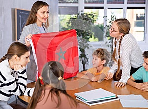 Group of curious preteen learners and young female teacher with flag of Morocco in classroom of academy