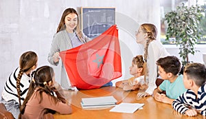 Group of curious preteen learners and young female teacher with flag of Morocco in classroom of academy