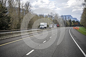 Group of different big rigs semi trucks with loaded semi trailers running on the turning wide highway road with winter bare trees