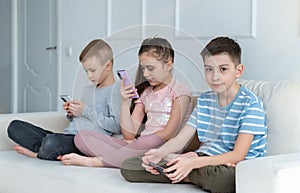 A group of different children are sitting in a row on the couch and emotionlessly playing online games or reading social networks.
