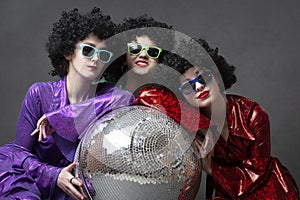 A group of disco girls in wigs with a disco ball and colorful costumes pose against a gray background