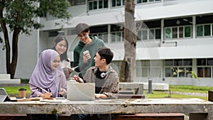 A group of diverse college students is discussing project details, using a laptop in a campus park