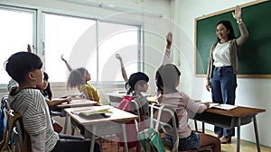 Group of diverse elementary school kids with notebooks sitting in classroom and raise arm up to answer teacher question. Education