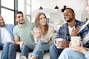 Group of diverse friends spending time togeher on weekend, holding hot drinks, having conversation and laughing at home