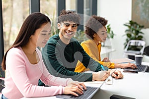 Group of diverse international students sitting at desk in coworking space, focus on smiling guy writing in copybook