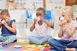 A group of diverse little girls having a tea party while playing on a colourful mat on the floor at preschool or