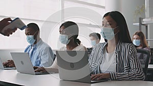 Group of diverse students in protective face masks typing on laptops, unrecognizable teacher measuring temperature