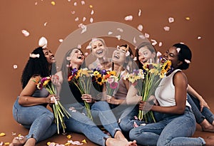 Group of diverse women with bouquets having fun under falling petals. Six happy females sitting together against a brown