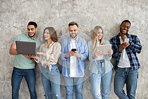 Group of diverse young friends with different gadgets studying or working together against grey studio wall