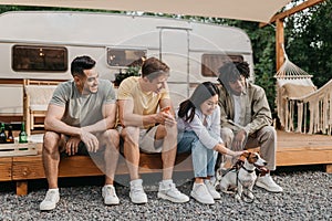 Group of diverse young friends sitting near camper van with pet dog, having fun together on camping trip