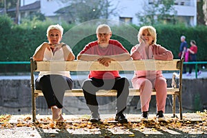 A group of elderly individuals, including a senior man and two older women, sits in a park on a sunny autumn day