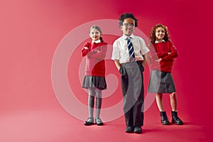 Group Of Elementary School Pupils Wearing Uniform Folding Arms Against Red Studio Background