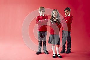 Group Of Elementary School Pupils Wearing Uniform Folding Arms Against Red Studio Background