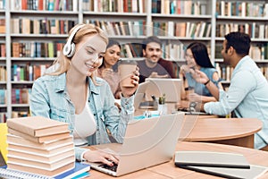 Group of ethnic multicultural students in library. White girl working on laptop with headphones.