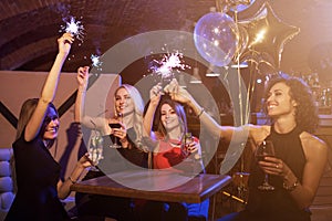 Group of female friends enjoying birthday party having fun with firework sparklers drinking alcoholic cocktails sitting