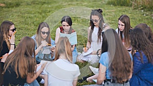 A group of female students are sitting in a circle on a meadow for collective work with notebooks.