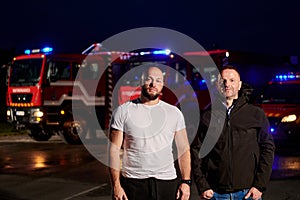 Group of firefighters, dressed in civilian clothing, stand in front of fire trucks during the night, showcasing a moment